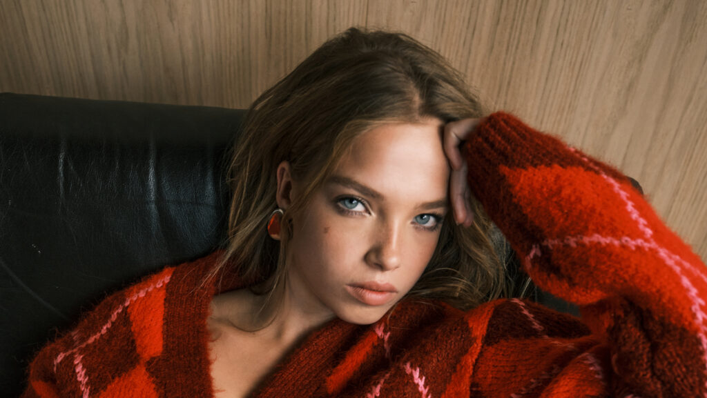 ASOS model in a red and pink argyle knit cardigan resting against a leather sofa