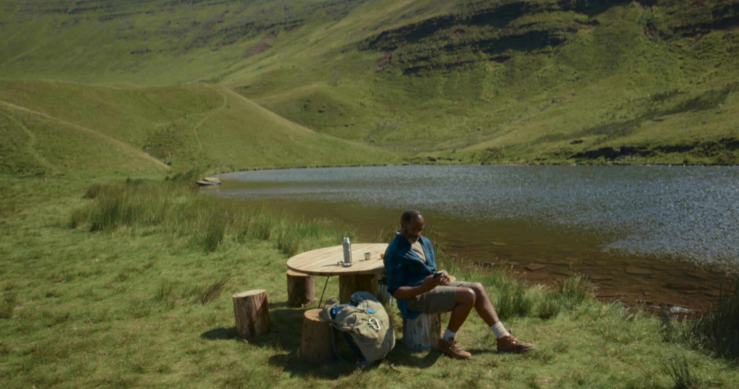 Man using a smartphone at a wooden table beside a lake in a remote green valley from the O2 satellite campaign