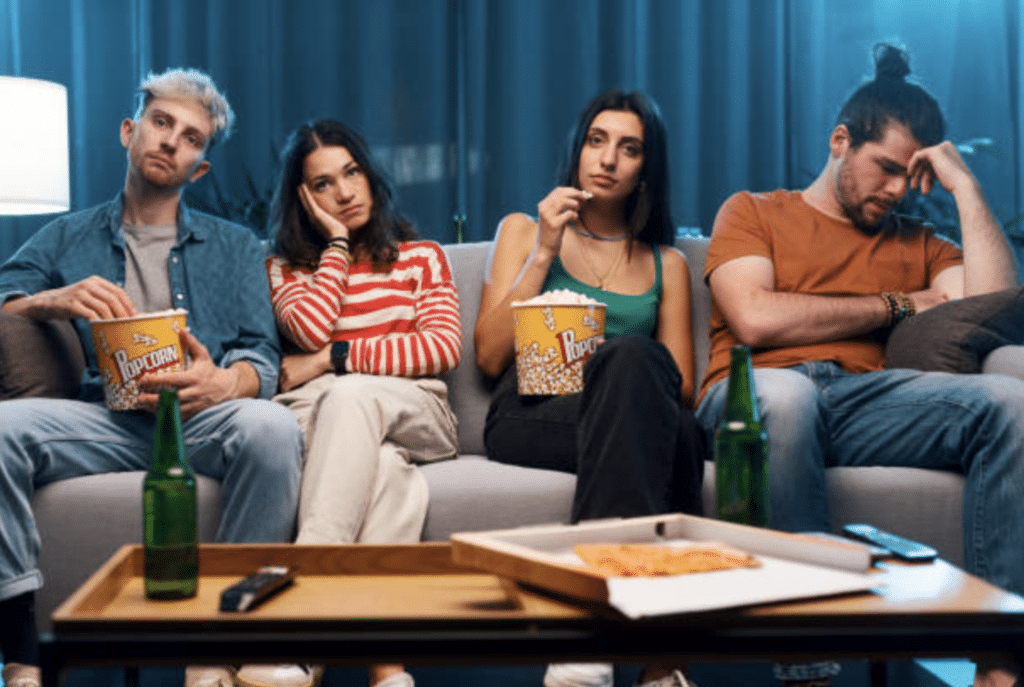 Four adults slumped together on a sofa with popcorn and snacks, lit by the glow of a screen, the kind of passive habit Virgin Media O2 says adds up to five years on their phones