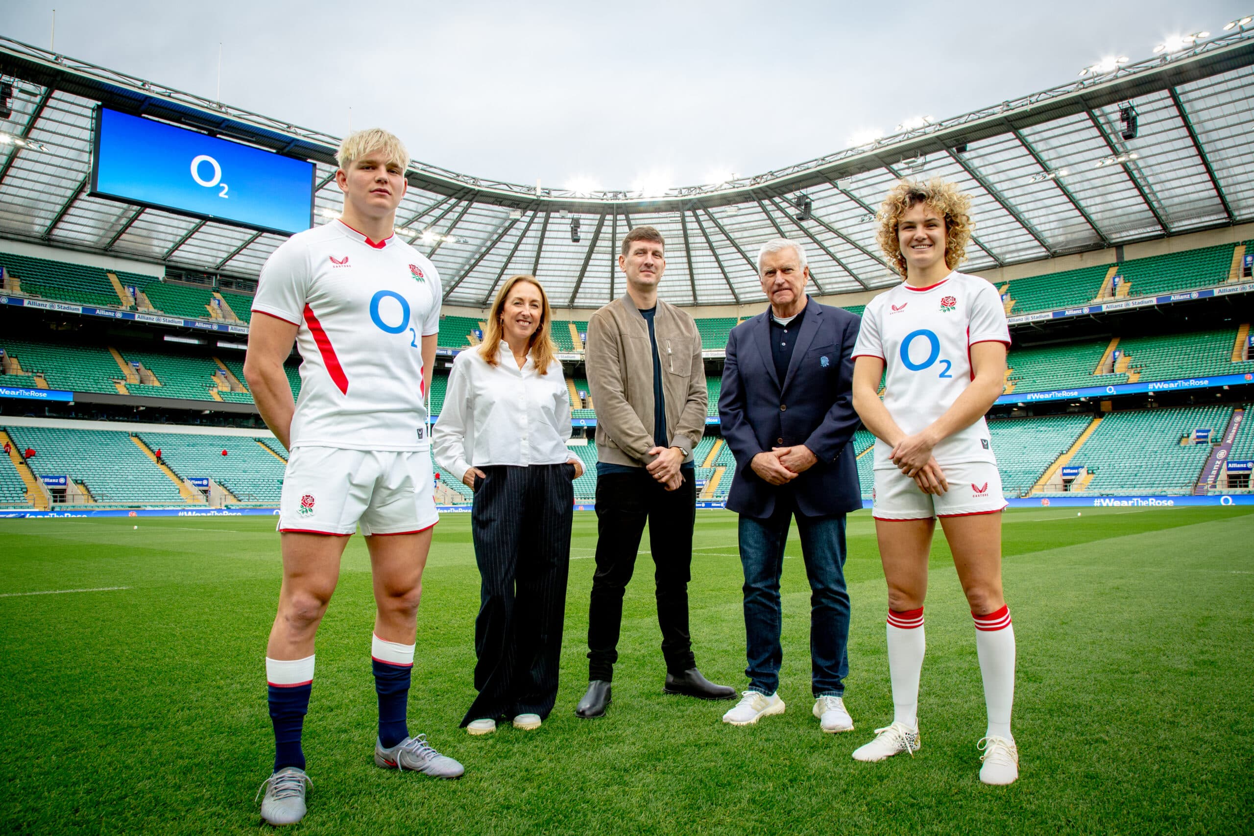 Two England rugby players in O2-branded shirts with executives on the pitch at Twickenham Stadium