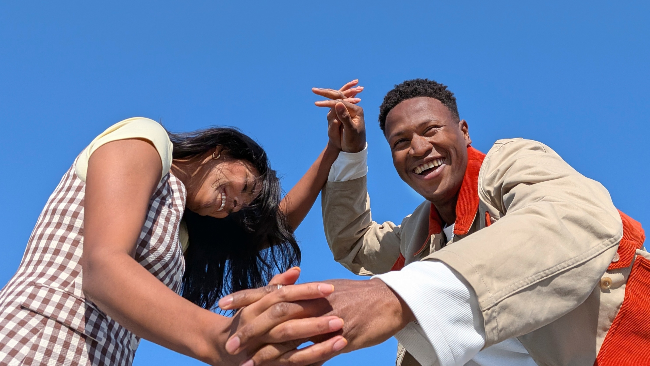 Two friends clasping hands and laughing against a clear blue sky, a photo shot on the current Pixel 10