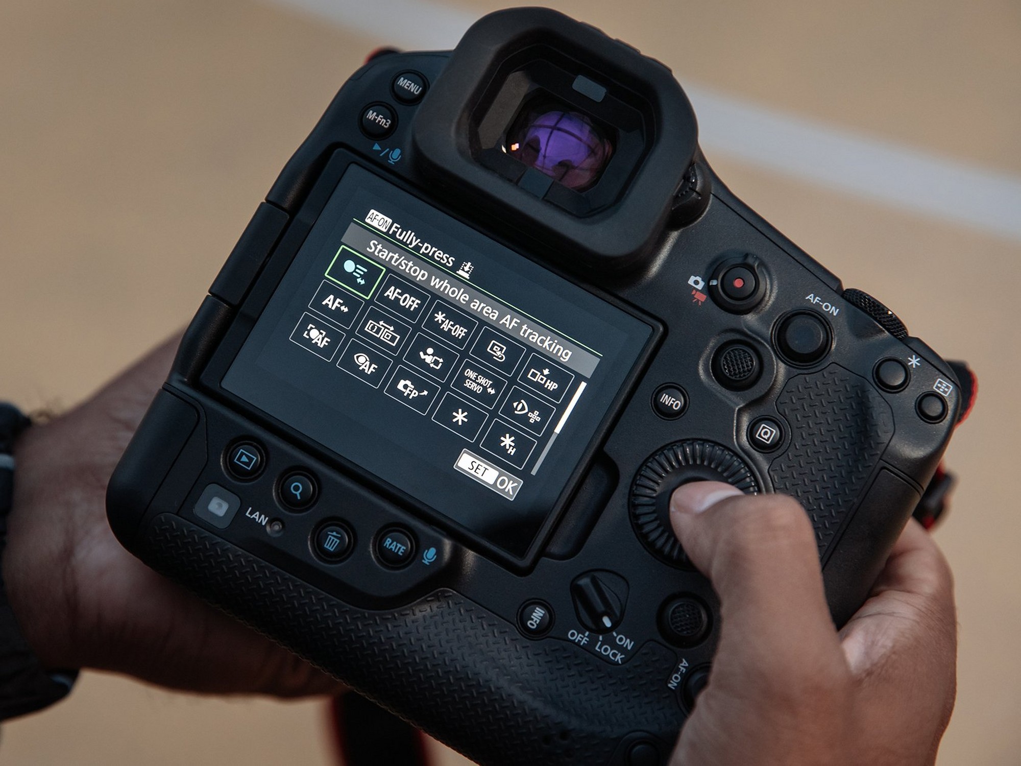 Hands holding the Canon EOS R1 showing the rear touchscreen with whole-area AF tracking menu