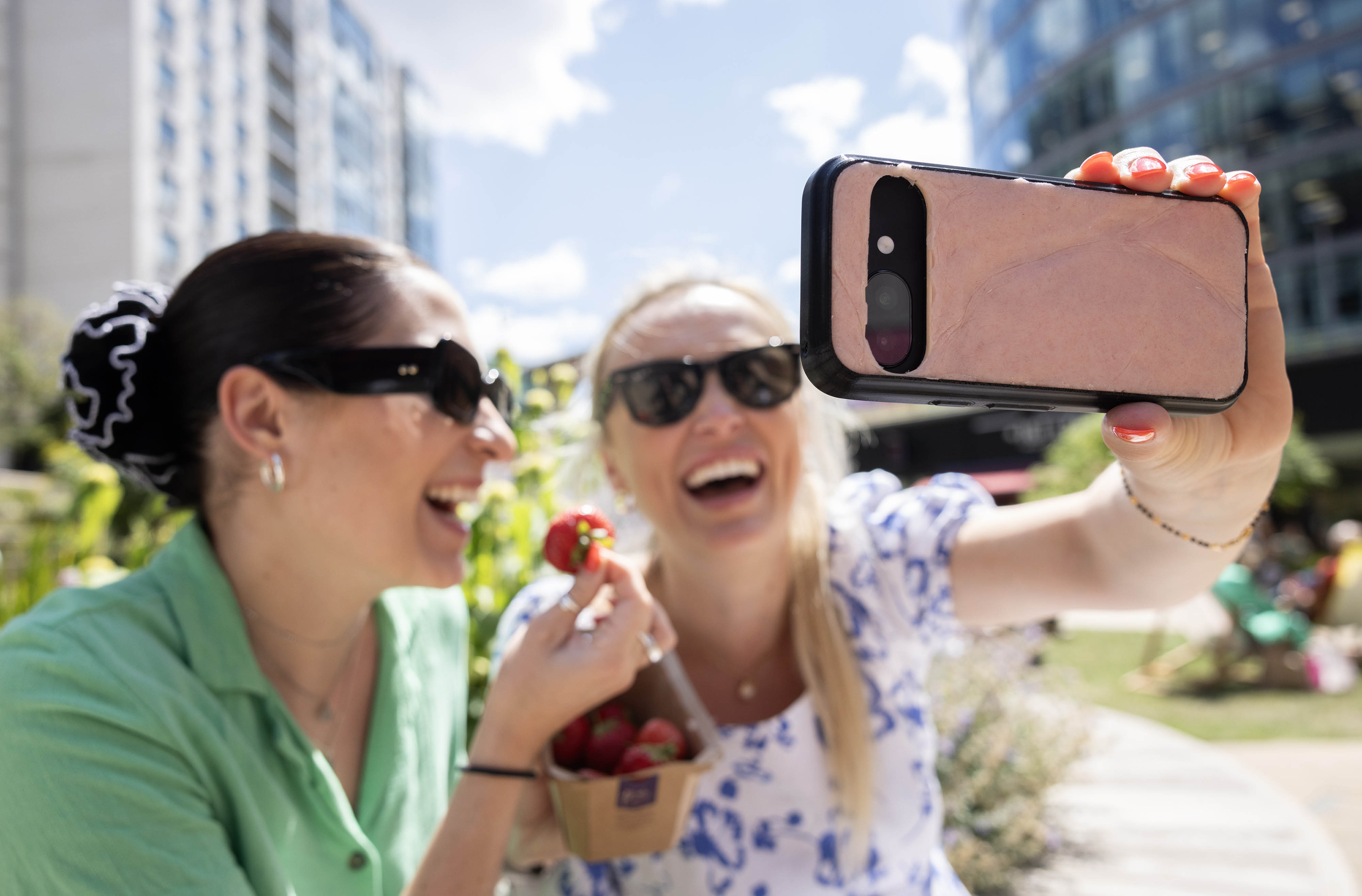 Two laughing women in sunglasses take a selfie on a smartphone in a sunny city garden, one holding a punnet of strawberries