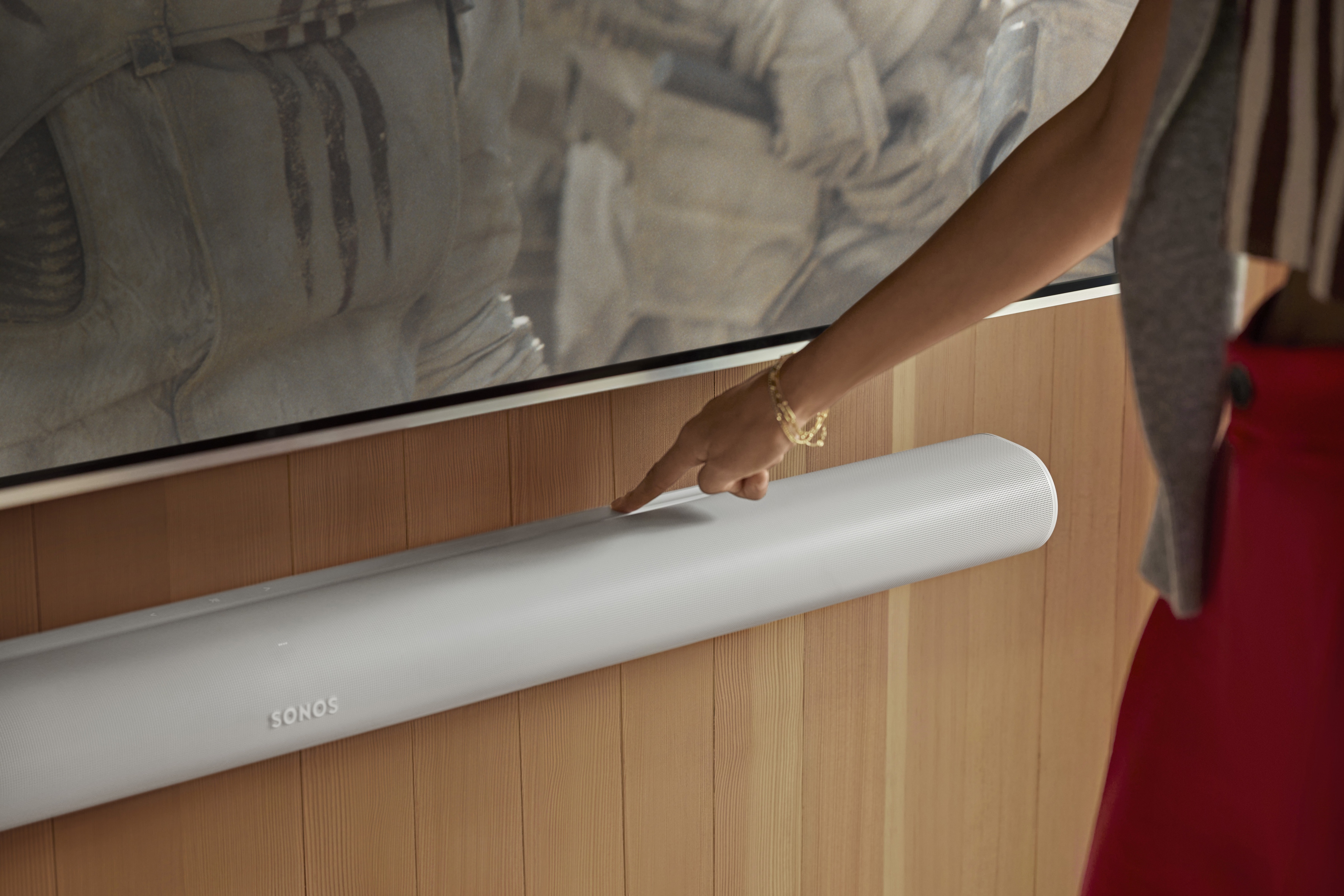 A person pressing the touch ledge of a white Sonos Arc Ultra mounted below a TV on a wood-panelled wall