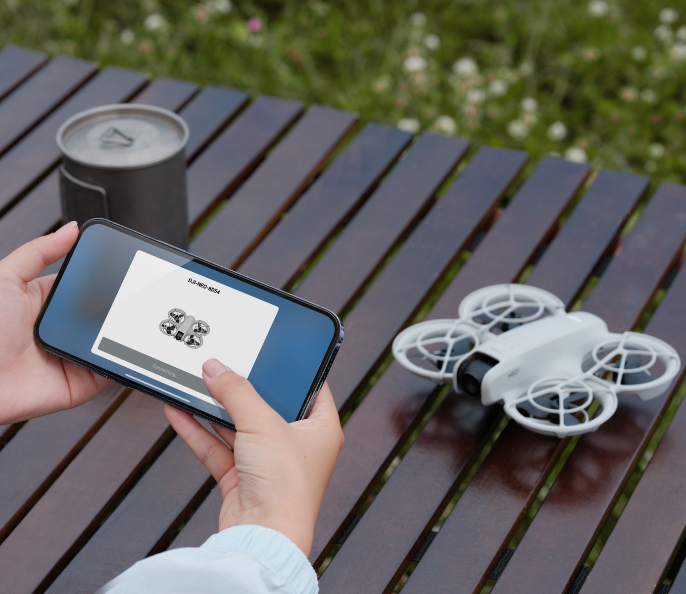 White DJI Neo drone with propeller guards on a wooden table next to a phone running the DJI app