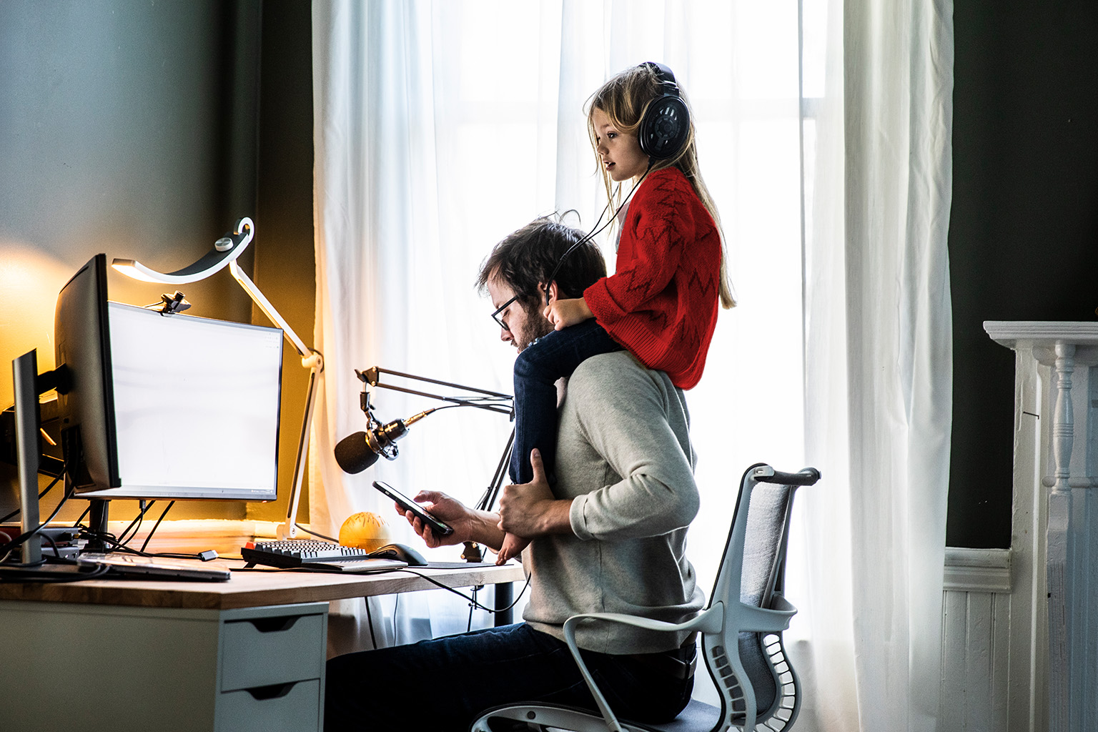 Father at a home-office desk holding a phone while his daughter sits on his shoulders wearing headphones