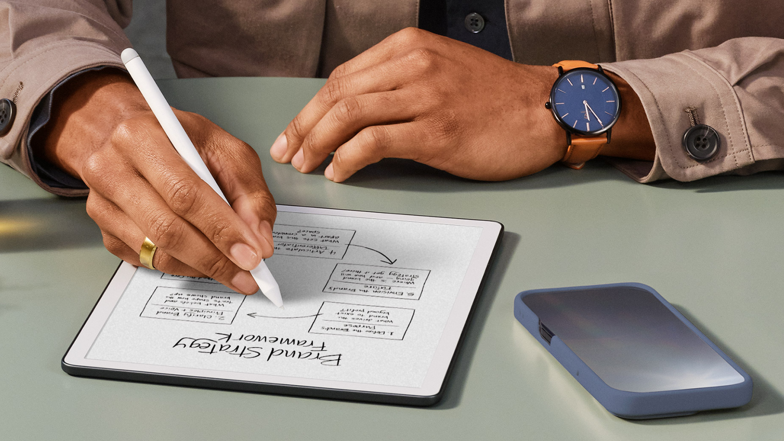 A person writing on a large-screen Kindle with the stylus, three-quarter view on a desk