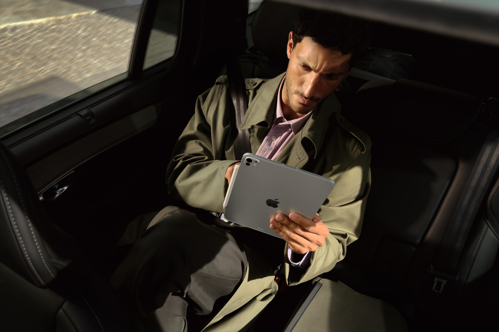 Man in a green coat reading on a space black iPad Pro M5 held in one hand in a sunlit car back seat