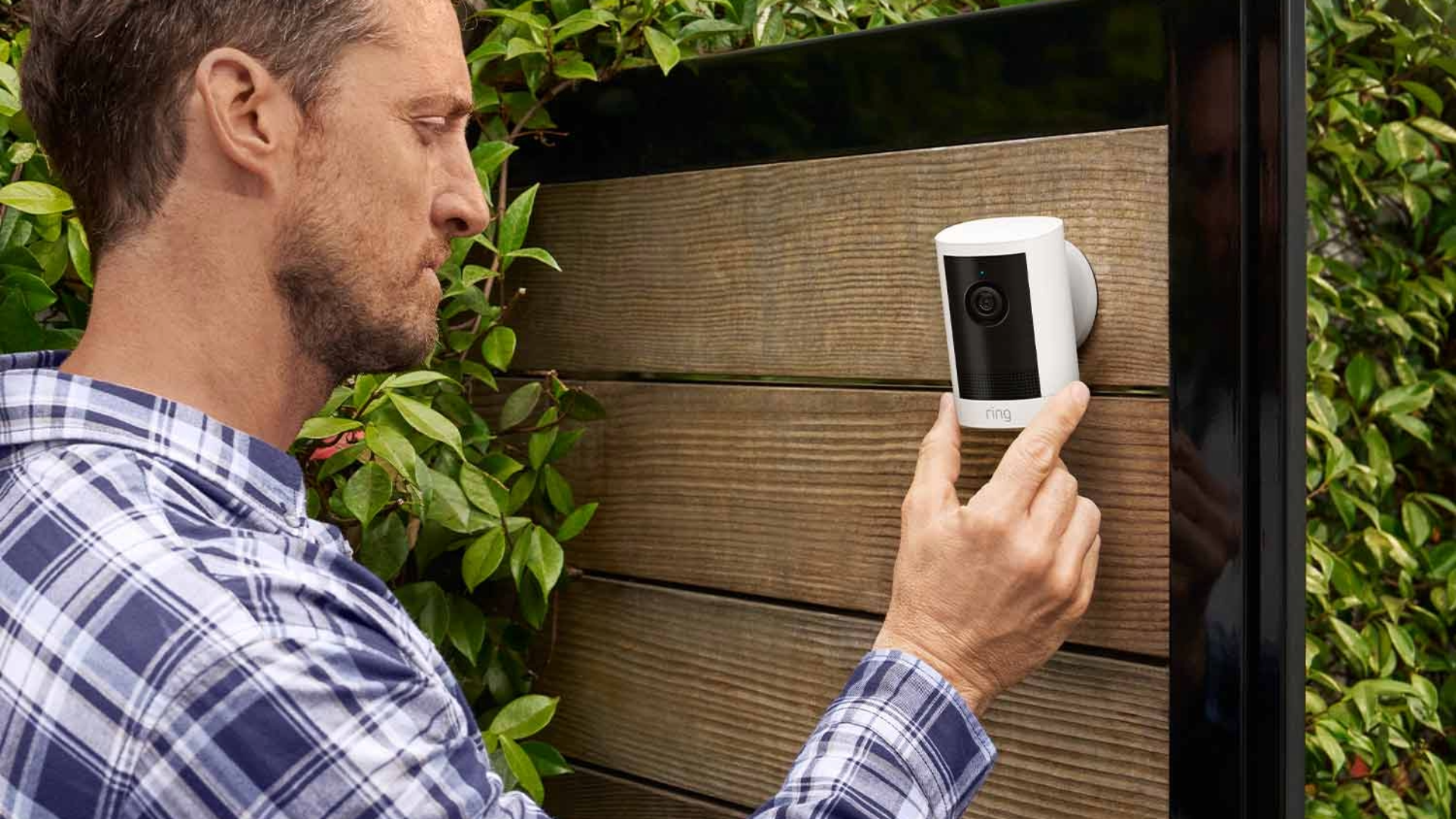 Ring outdoor security camera in white held in a person's hand against wooden fencing