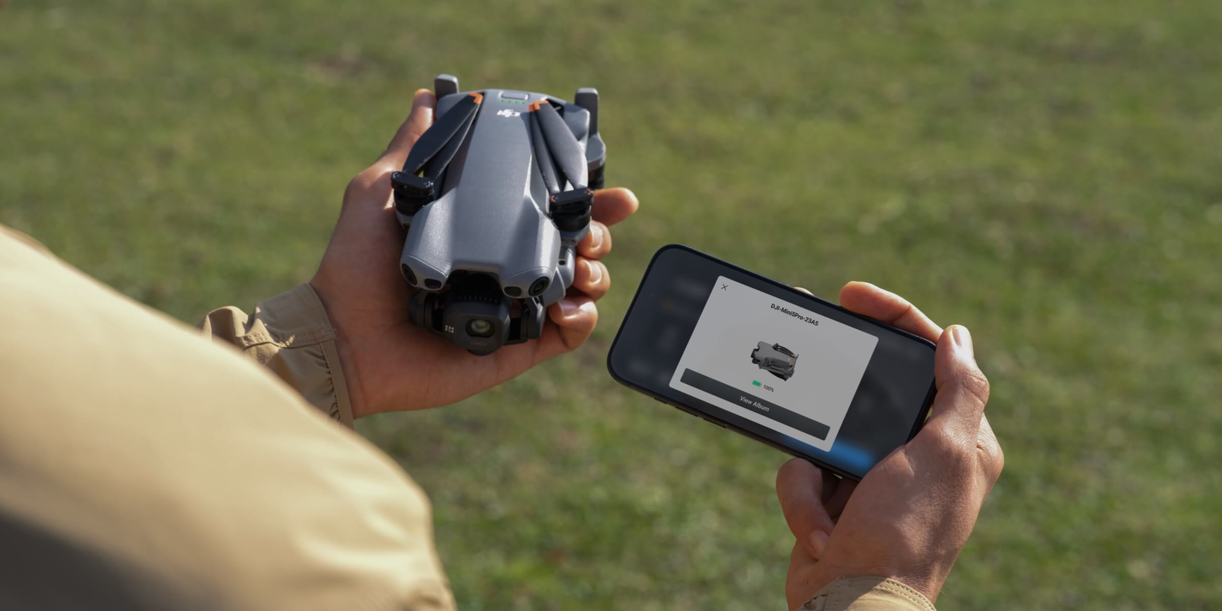DJI Mini 5 Pro folded in a hand next to a phone showing the DJI Fly app at 100 percent